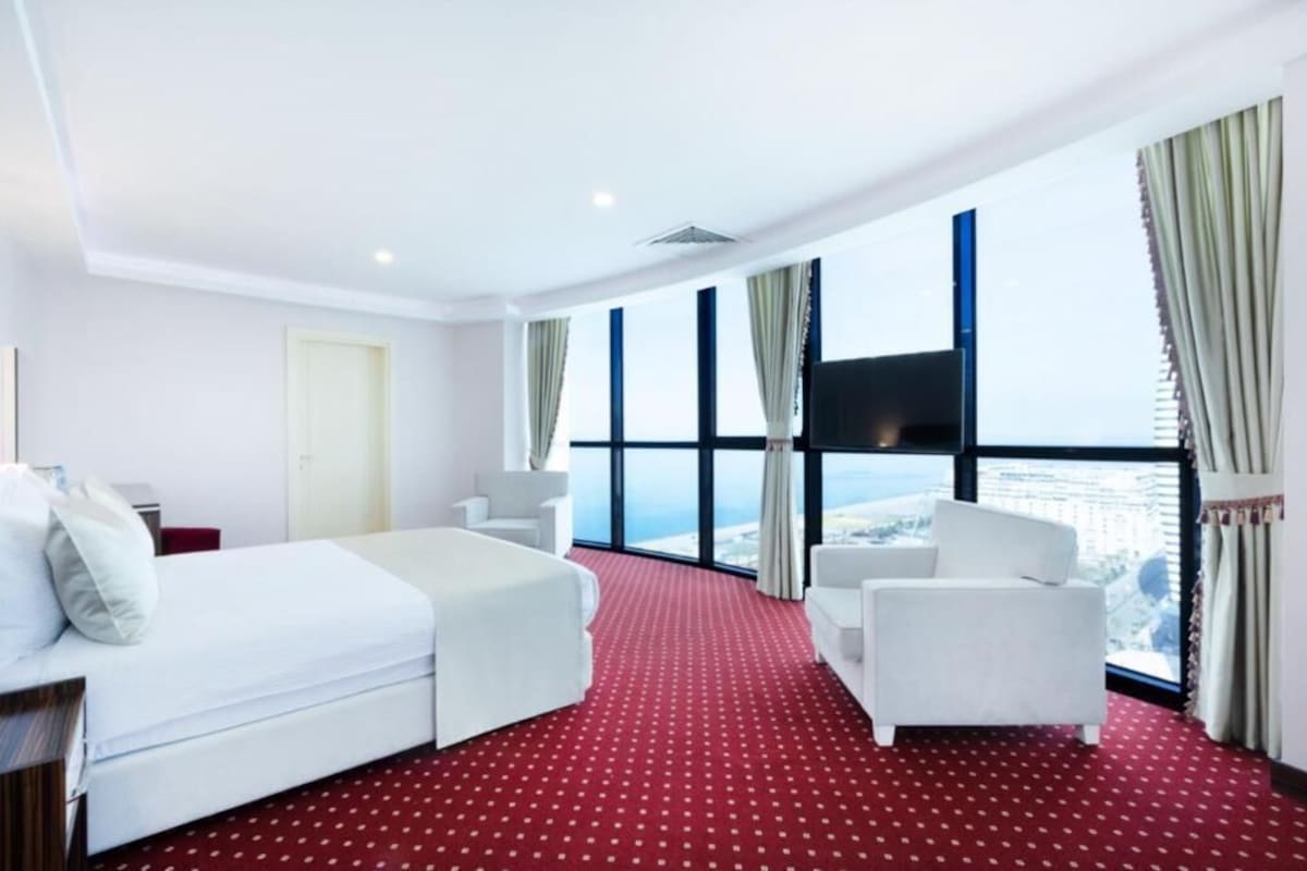 A hotel room with a bed, two chairs, a desk, and a large window offering a city view.