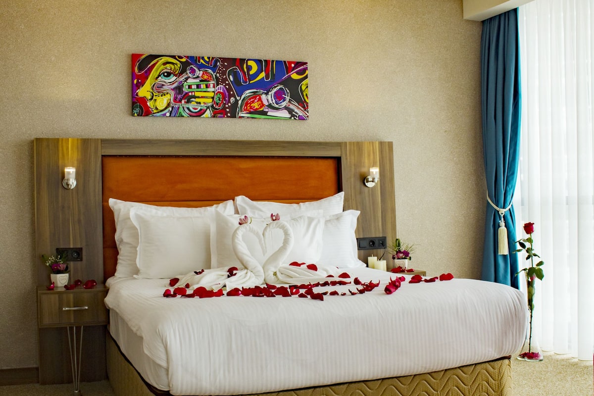 A hotel room with a large bed, a headboard, bedside tables, a painting on the wall, and a window with curtains.