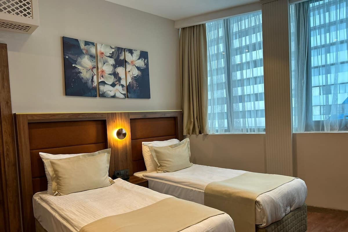 A hotel room with two beds, a headboard with a circular light, a painting on the wall, and a window with curtains.