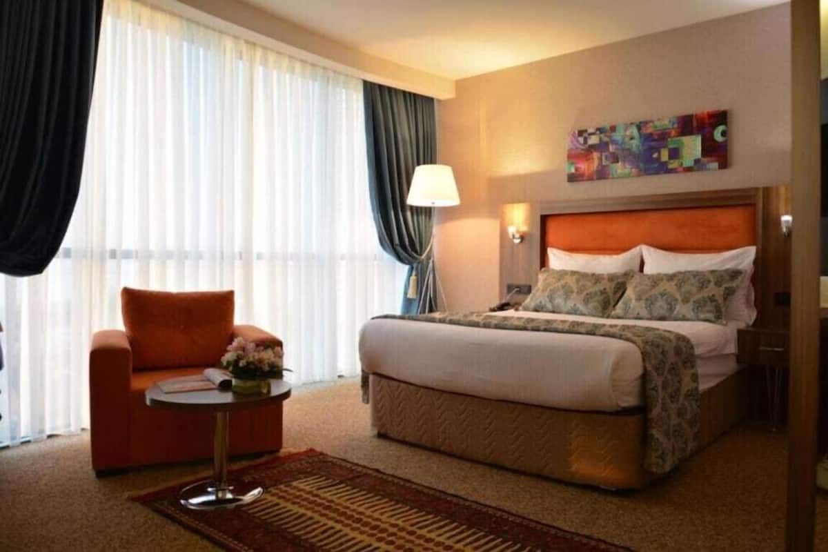 A hotel room with a bed, a chair, a table, a lamp, and a painting on the wall.