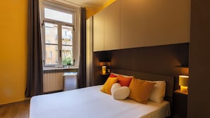 Ein Hotelzimmer mit einem großen Bett, einem Fenster mit Vorhängen, einem Nachttisch mit Lampe und Blick auf Gebäude.