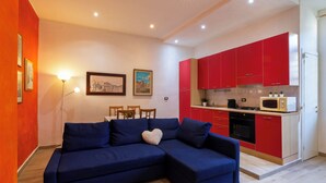 Ein Wohnzimmer mit einem blauen Ecksofa, einem Esstisch mit Stühlen und einer Küche mit roten Schränken und einer Mikrowelle.
