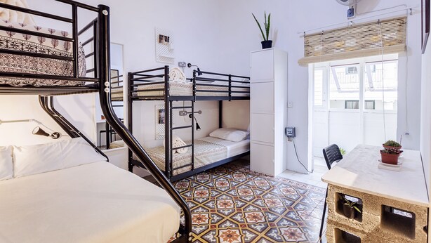 A compact room with bunk beds, a desk, a potted plant, and a window with blinds. - Granny's Inn - Hostel (Sliema)