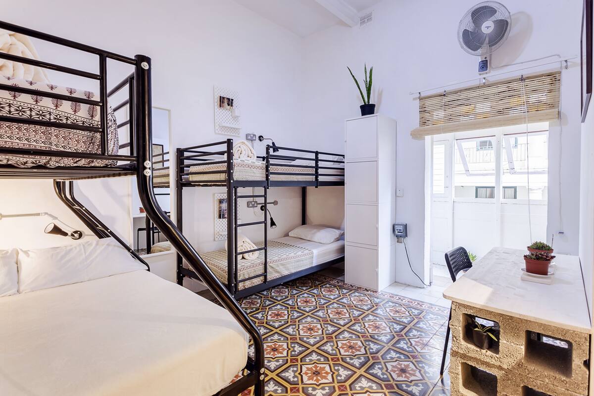 Quarto compacto com beliches, escrivaninha, planta em vaso e janela com persianas.