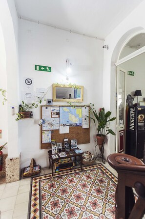 Una habitación acogedora con un tablón de corcho lleno de mapas y notas, una cafetera Nescafé, una planta en maceta y una alfombra estampada.