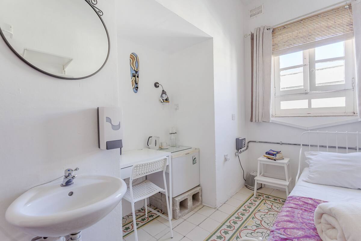 Quarto pequeno e limpo com cama, pia branca, espelho, mesa pequena e janela com persianas.