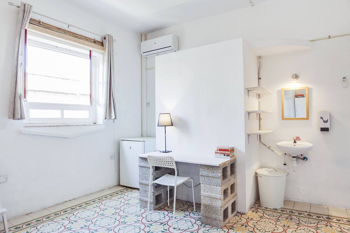 Quarto compacto com escrivaninha, cadeira, janela, ar condicionado, pia e piso cerâmico.