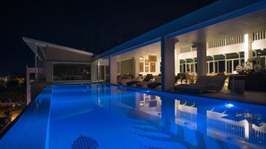 Una piscina infinita con borde azul cristalino, un área de estar exterior moderna con sillones de descanso, y un espacio interior bien iluminado con zona de comedor.