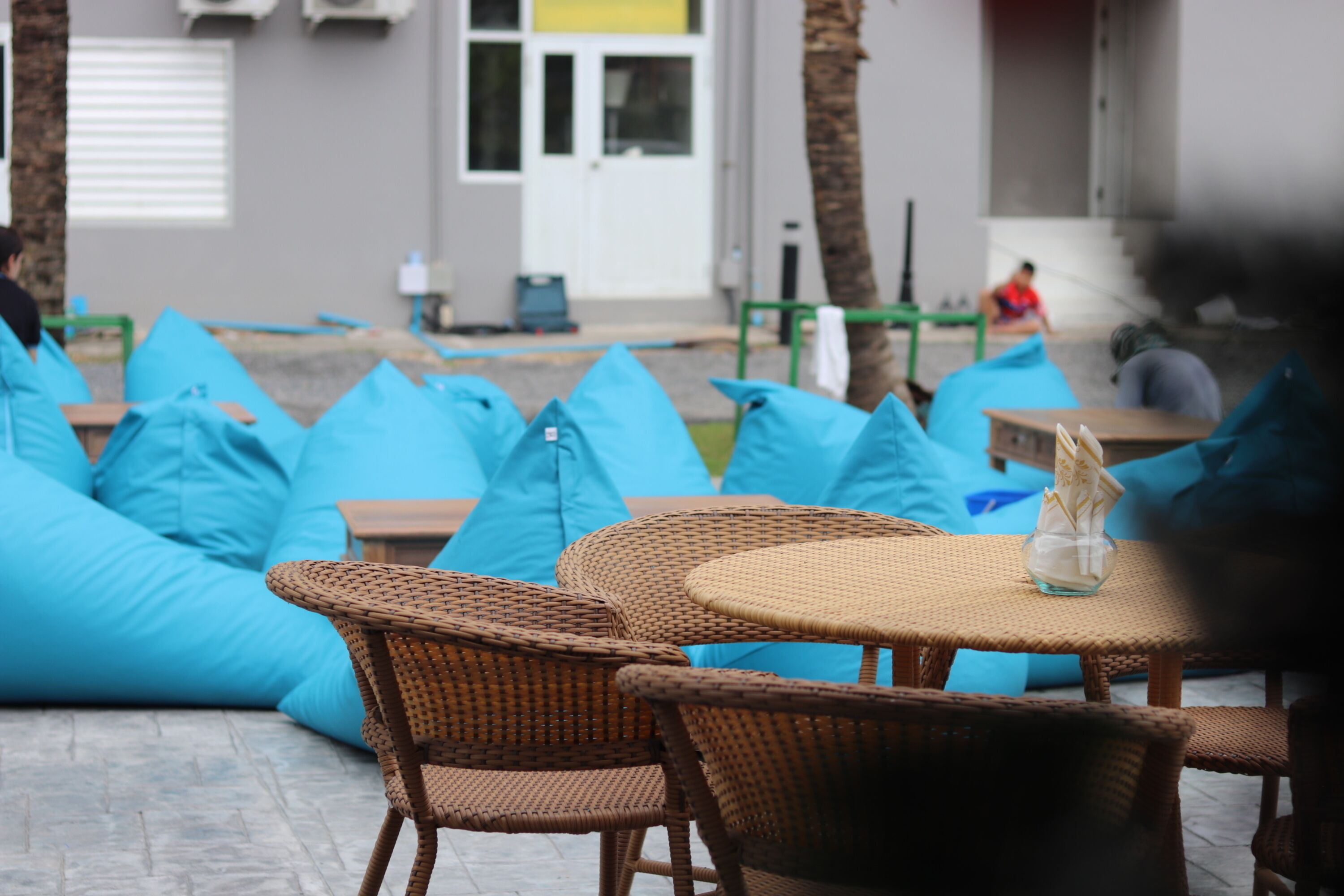 Ein Terrassenbereich mit Korbstühlen und Tischen, blauen Kissen und einer Glasvase auf einem der Tische.