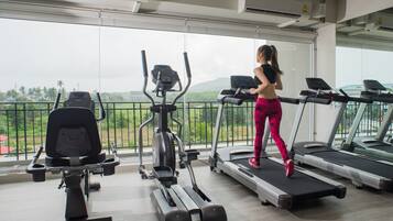 A gym with treadmills, an elliptical, and a rowing machine, with a view of greenery and mountains through large windows.