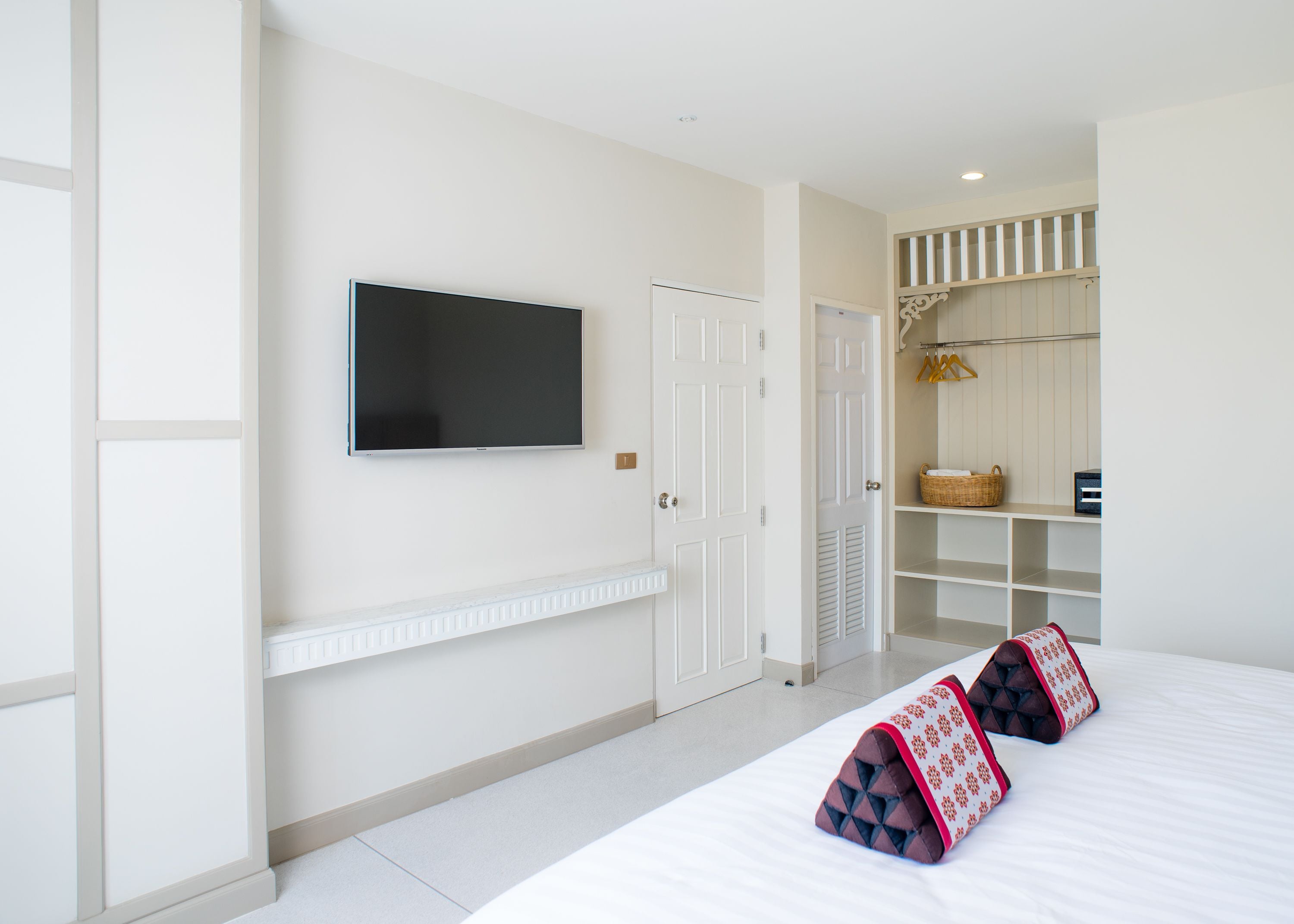 Een hotelkamer met een bed, een flatscreen televisie aan de muur, een witte deur en een kast met planken en een mand.