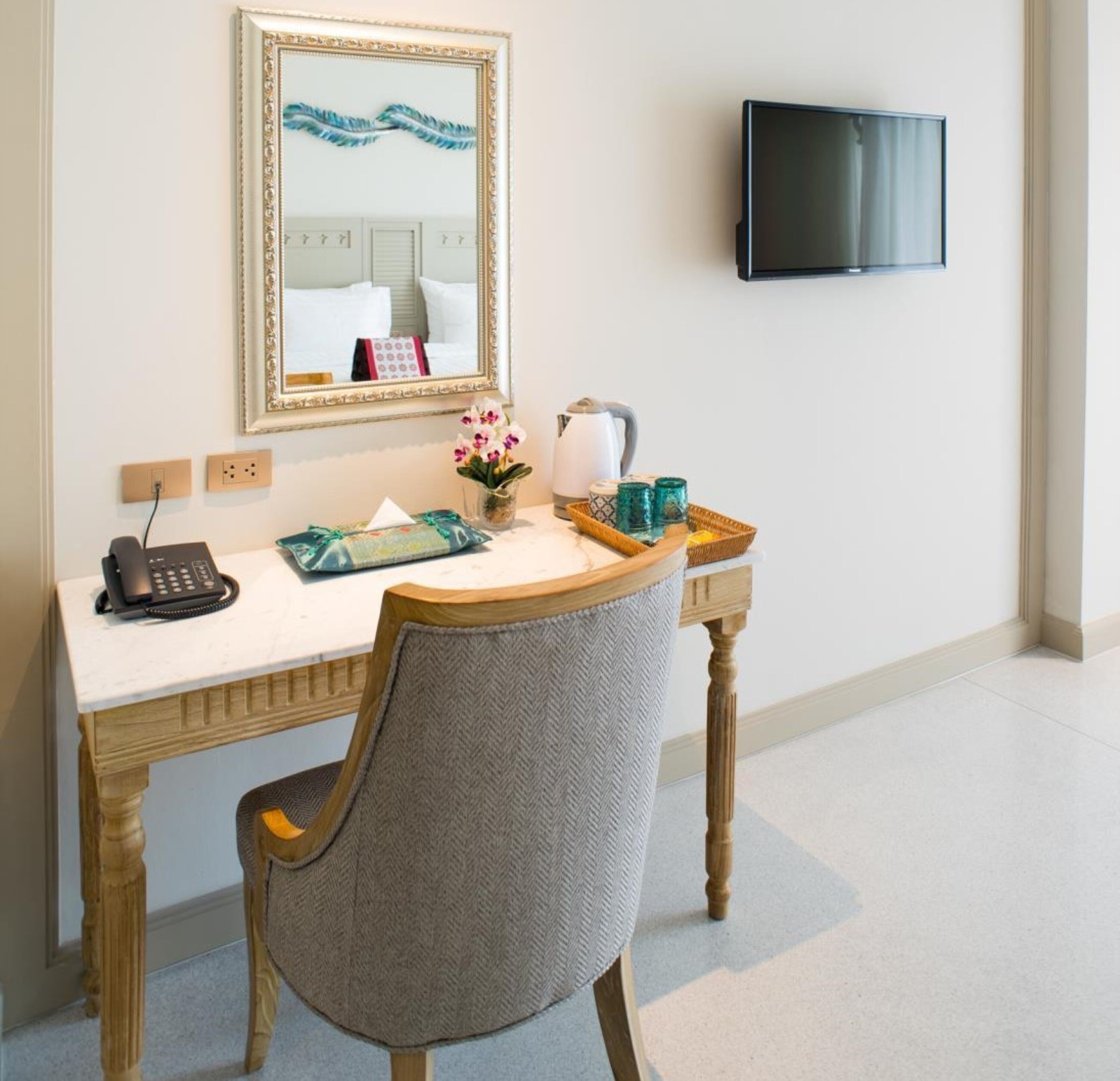A hotel room with a wooden desk, a chair, a telephone, a mirror, a television, and a small table with a kettle, cups, and a vase with flowers.