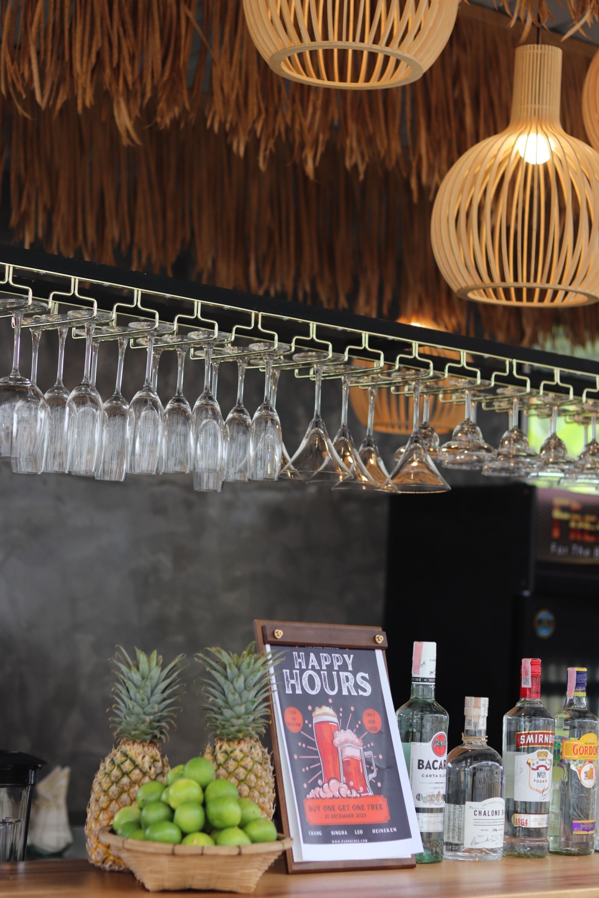 A bar area with a 'Happy Hours' sign, a selection of bottles, a basket of limes, and a row of hanging glasses.