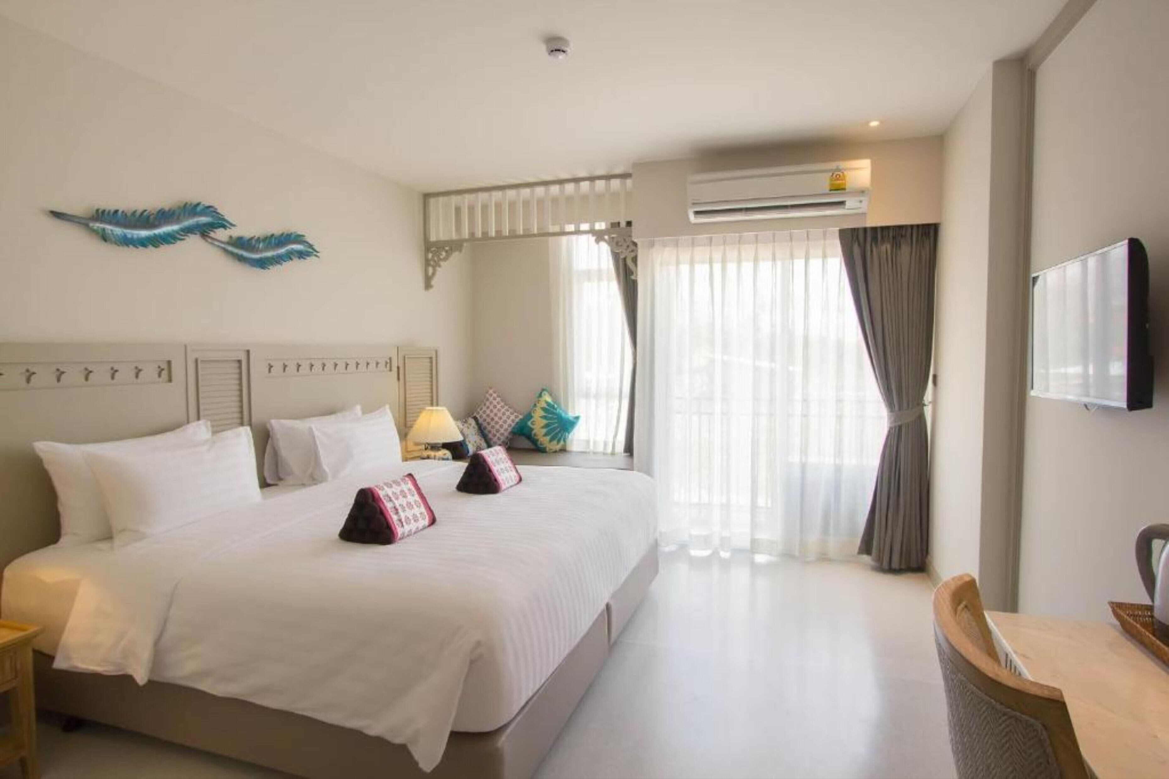 A hotel room with a large bed, a desk, a chair, an air conditioning unit, a television, and decorative pillows.