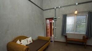 Une pièce comprenant un lit simple, un banc, une fenêtre avec des stores et une porte donnant sur une autre pièce.