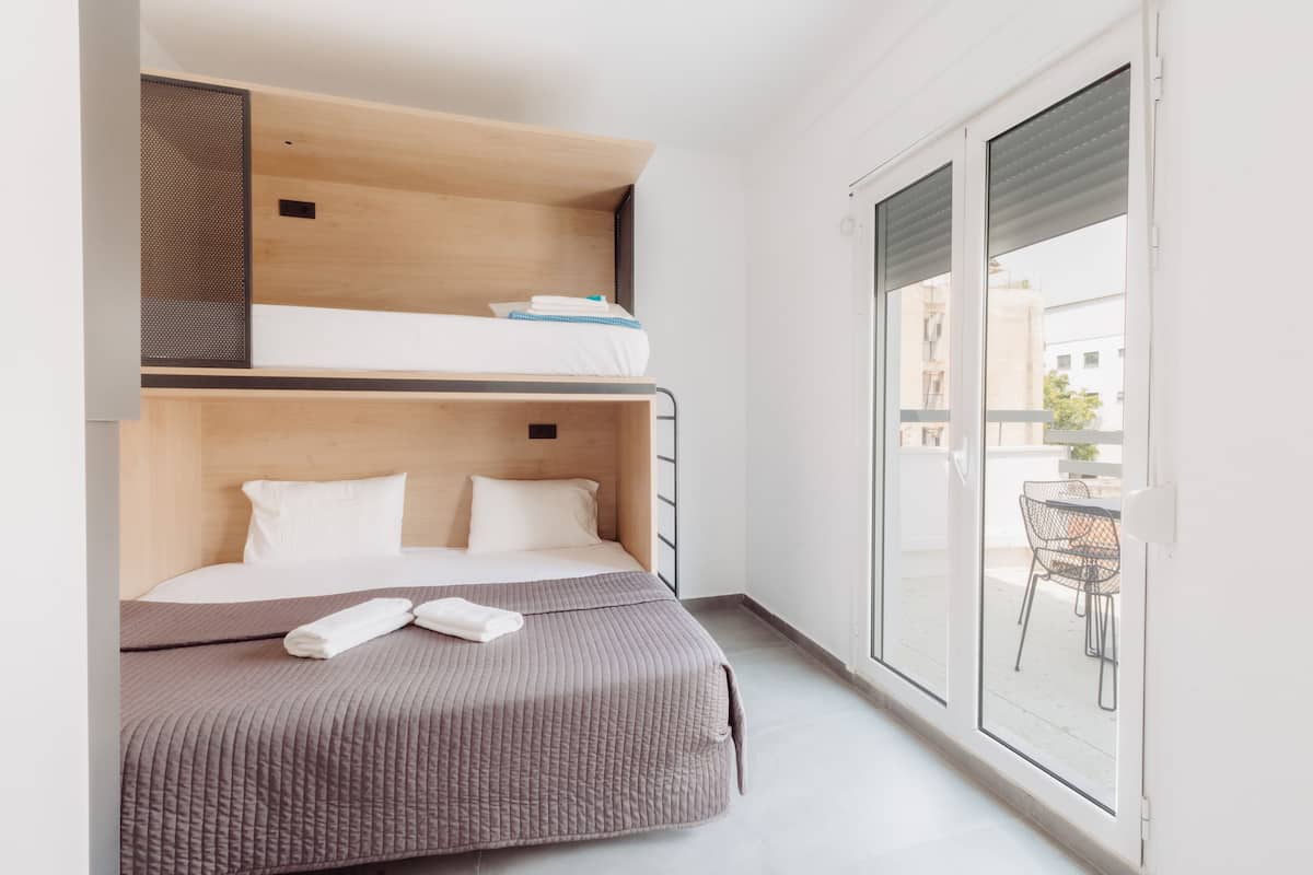 A modern hotel room with a bunk bed, a large window with a view of the city, and a balcony with outdoor seating.