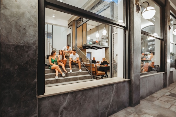 A modern interior with a staircase leading to an upper level, a seating area with tables, and a large window offering a view of the street. - Athens Backpackers (Athens)