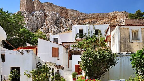 A narrow street with white buildings, a sign reading "ACROPOLIS," and a rocky hill in the background.