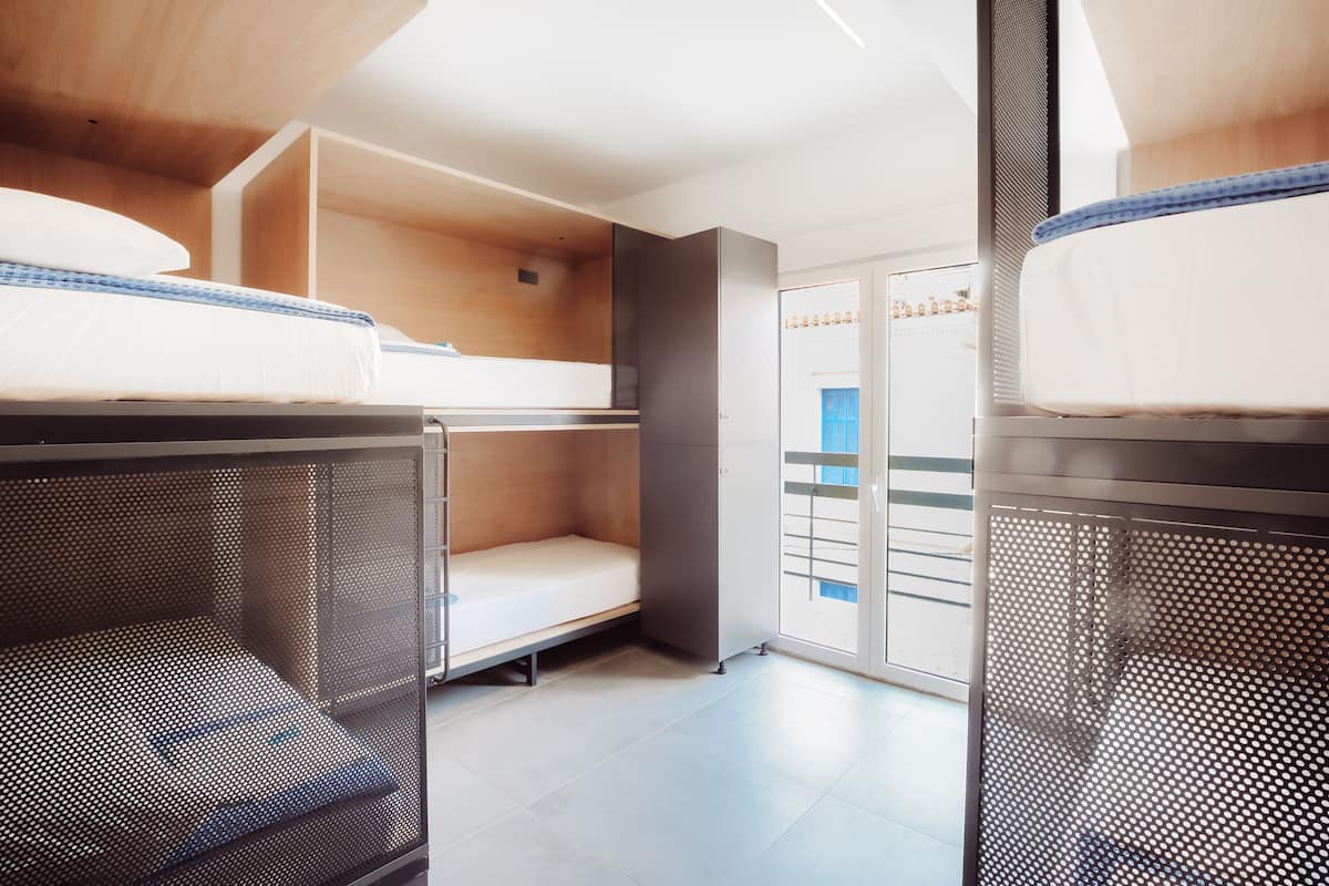 A dormitory room with bunk beds, a small kitchenette, and a sliding door leading to a balcony.