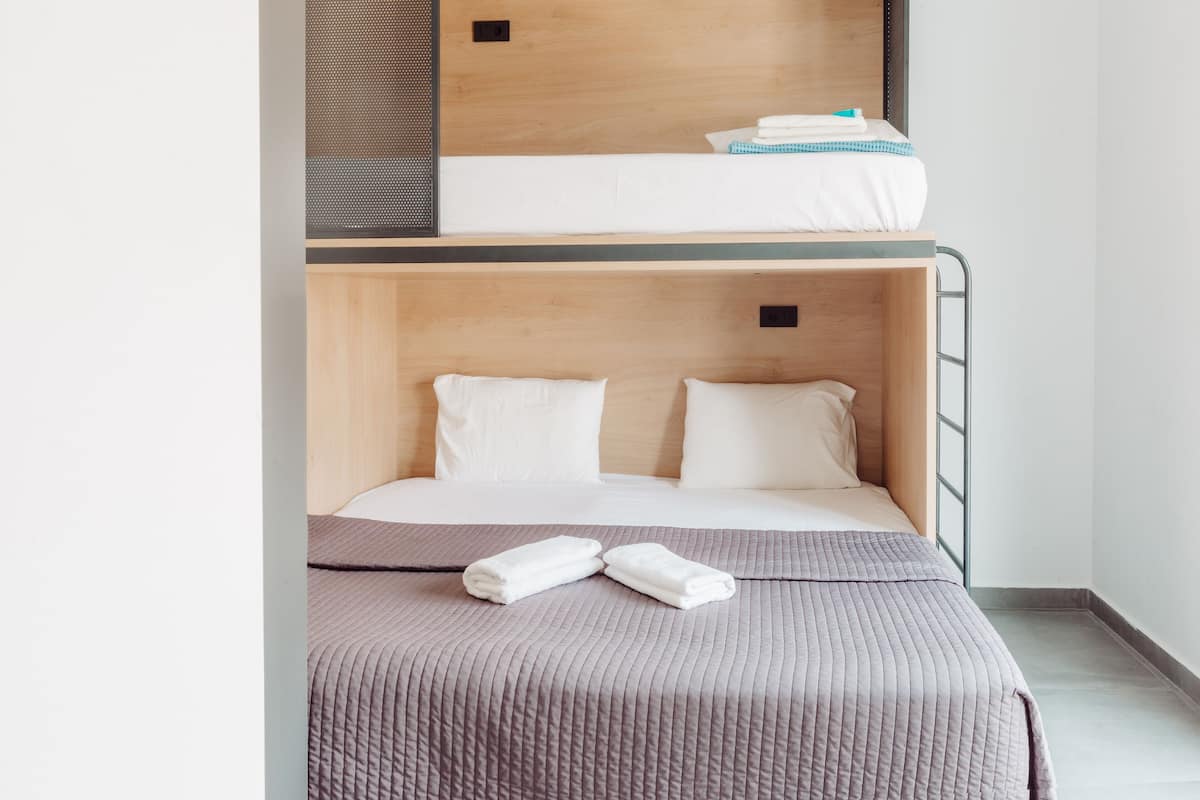 A bunk bed with a lower bed and a top bunk. The lower bed has a grey quilt and two white pillows. There are two folded towels on the lower bed. The top bunk has a white mattress with a blue and white checkered blanket.