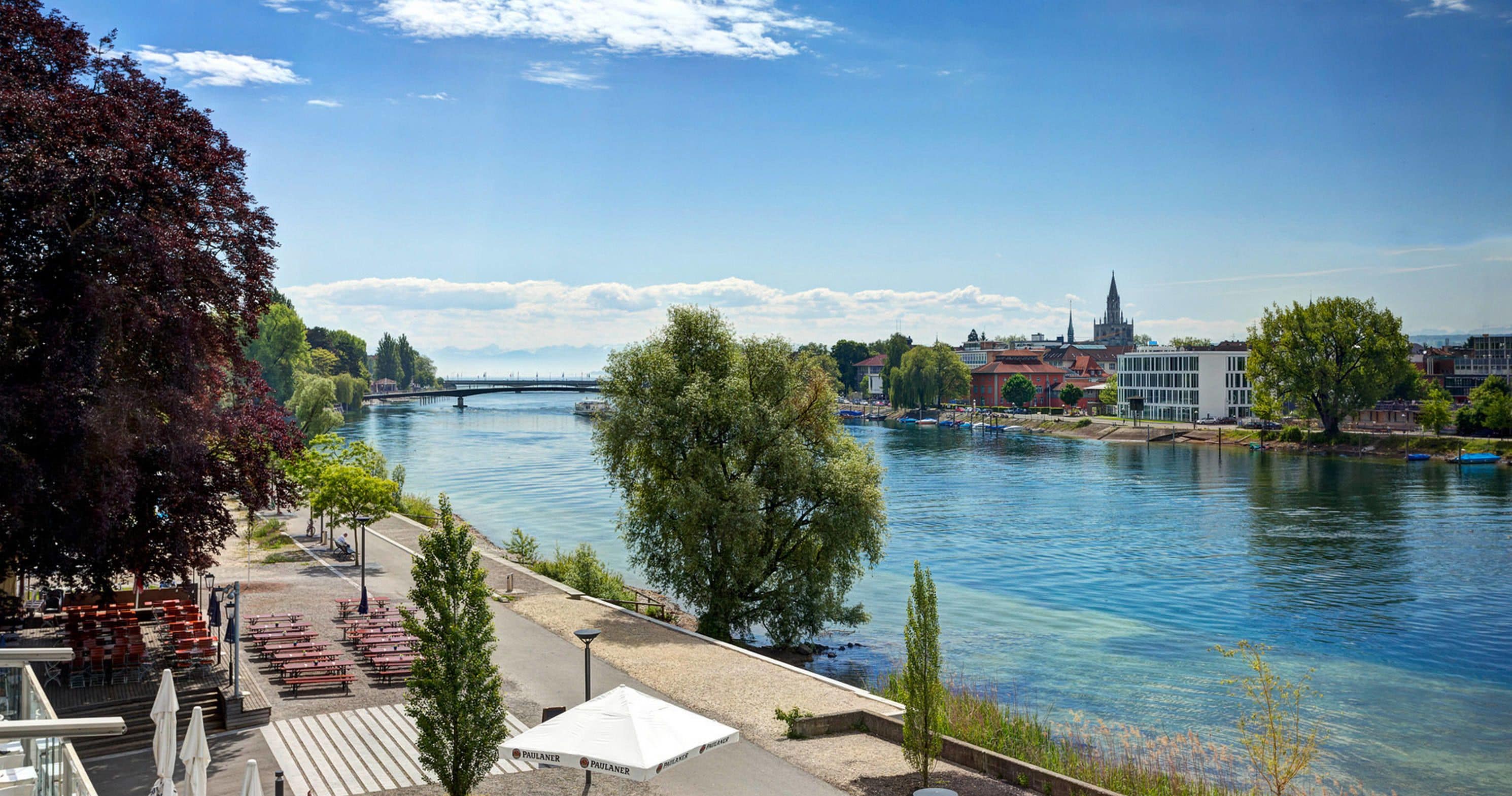 A riverside view with a bridge, dining area, and buildings in the background.