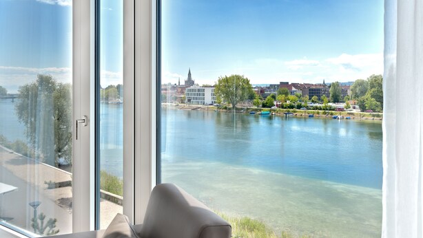 Een kamer met een groot raam met uitzicht op een rivier, een stoel en een beeld van gebouwen aan de overkant.