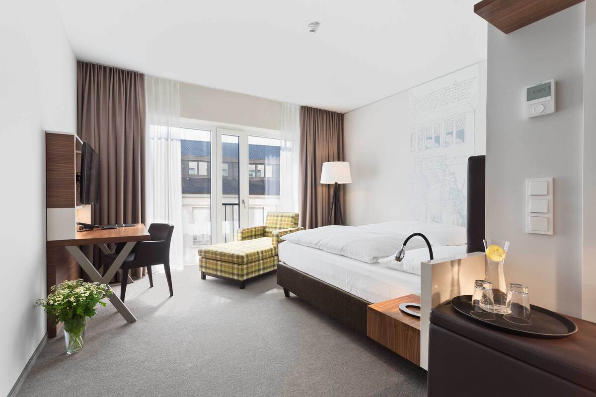 Una habitación de hotel con cama, escritorio, silla y ventana con cortinas.