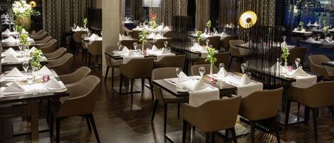 A restaurant with multiple tables set for dining, featuring a modern interior with wooden flooring and a view of the cityscape through large windows.