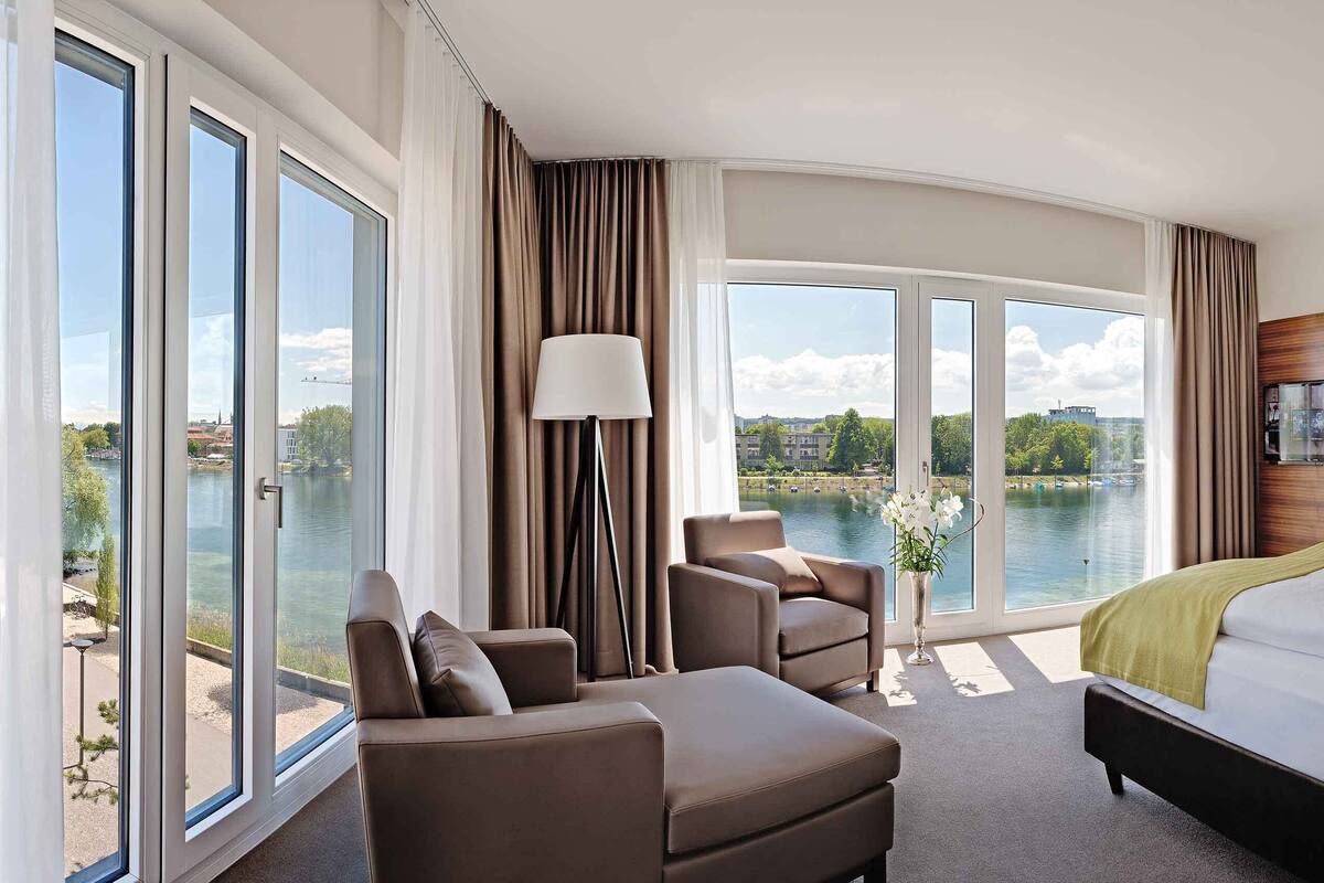 Una habitación de hotel moderna con una cama grande, un sofá y una silla, todos orientados a una vista panorámica de un lago a través de amplios ventanales.