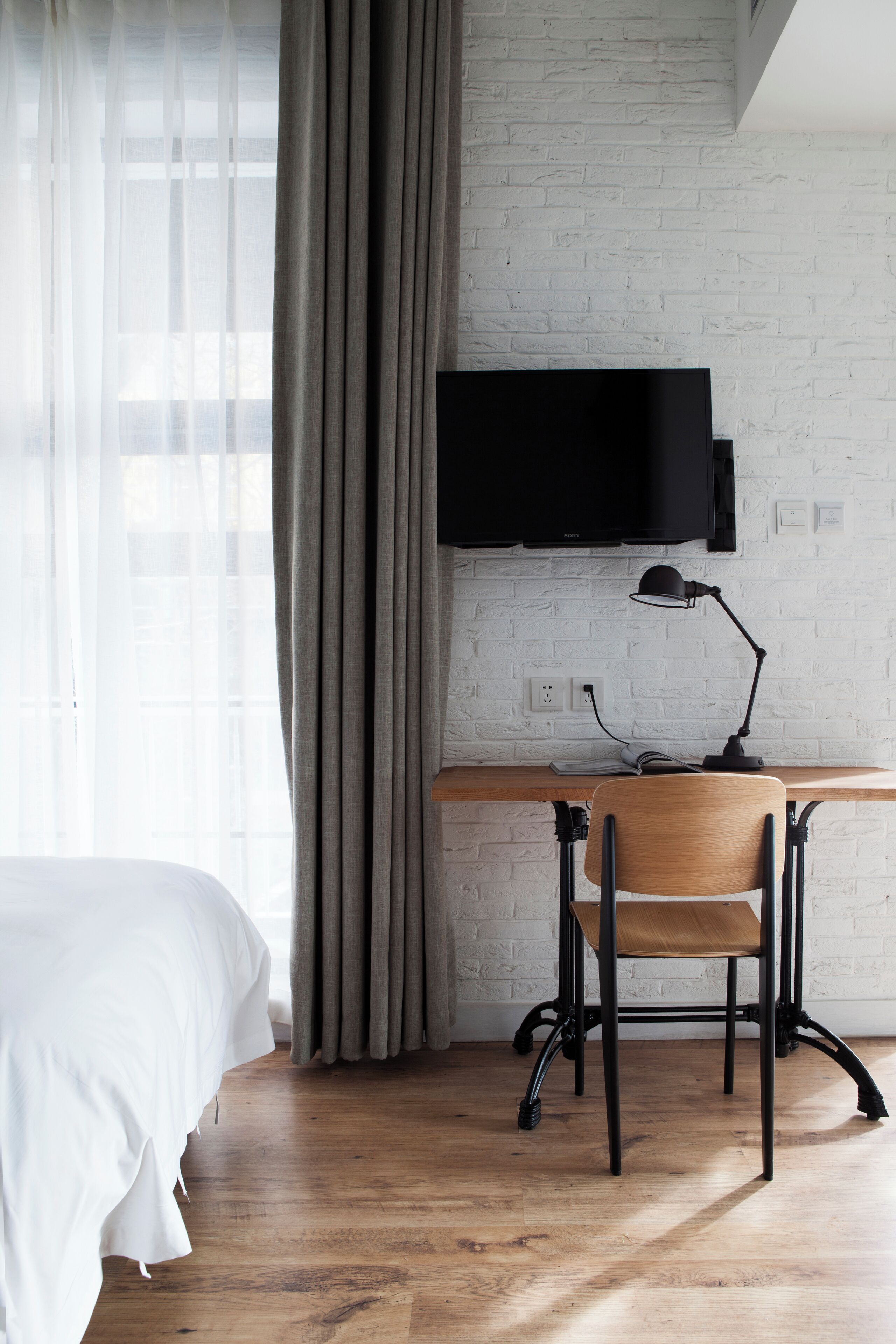A hotel room with a bed, a desk with a chair, a television mounted on a white brick wall, and a window with curtains.
