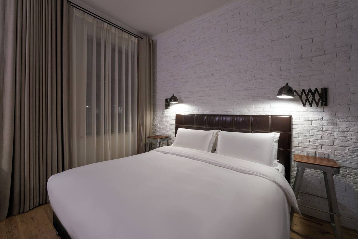 Una cama bien tendida en una habitación con pared de ladrillo blanco, dos mesitas de noche y una ventana con cortinas.
