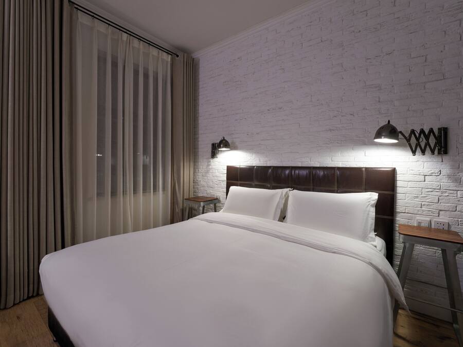 Cama bem arrumada em quarto com parede de tijolos brancos, duas mesas de cabeceira e janela com cortinas.
