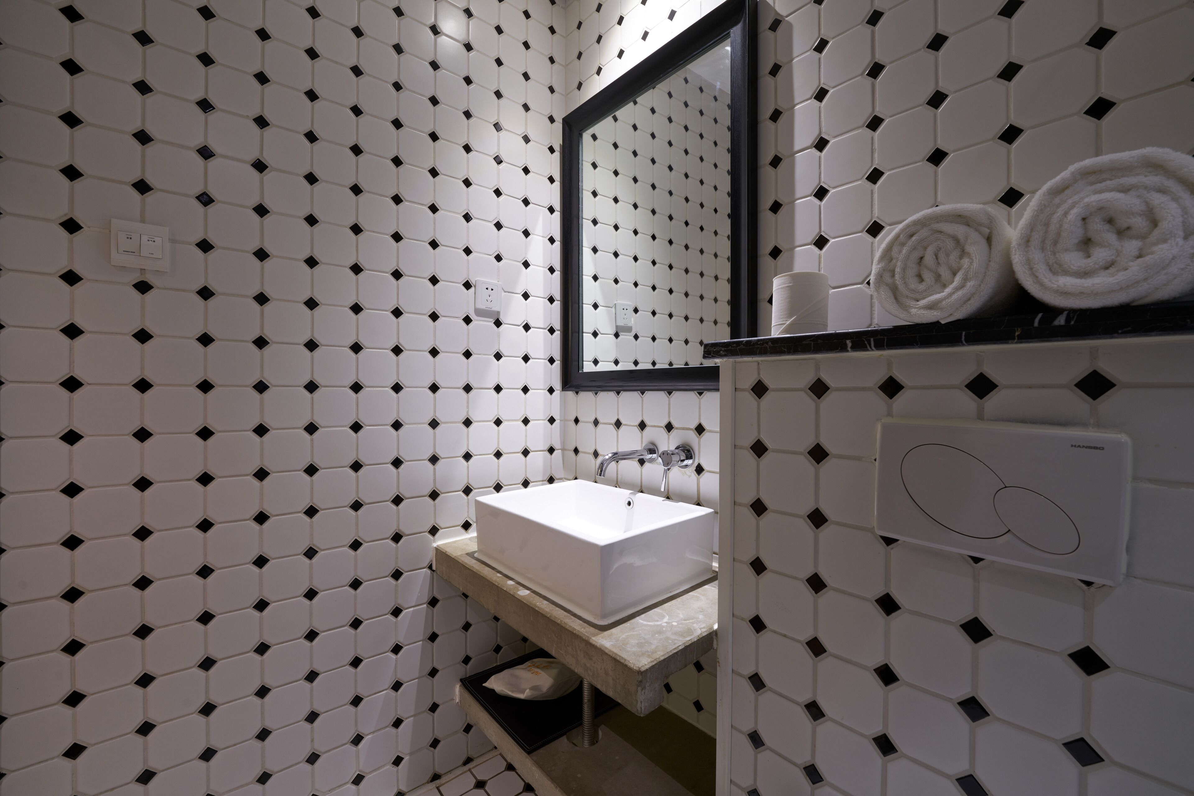 A bathroom with a white sink, black diamond-patterned tiles, a mirror, and two rolled towels on the counter.
