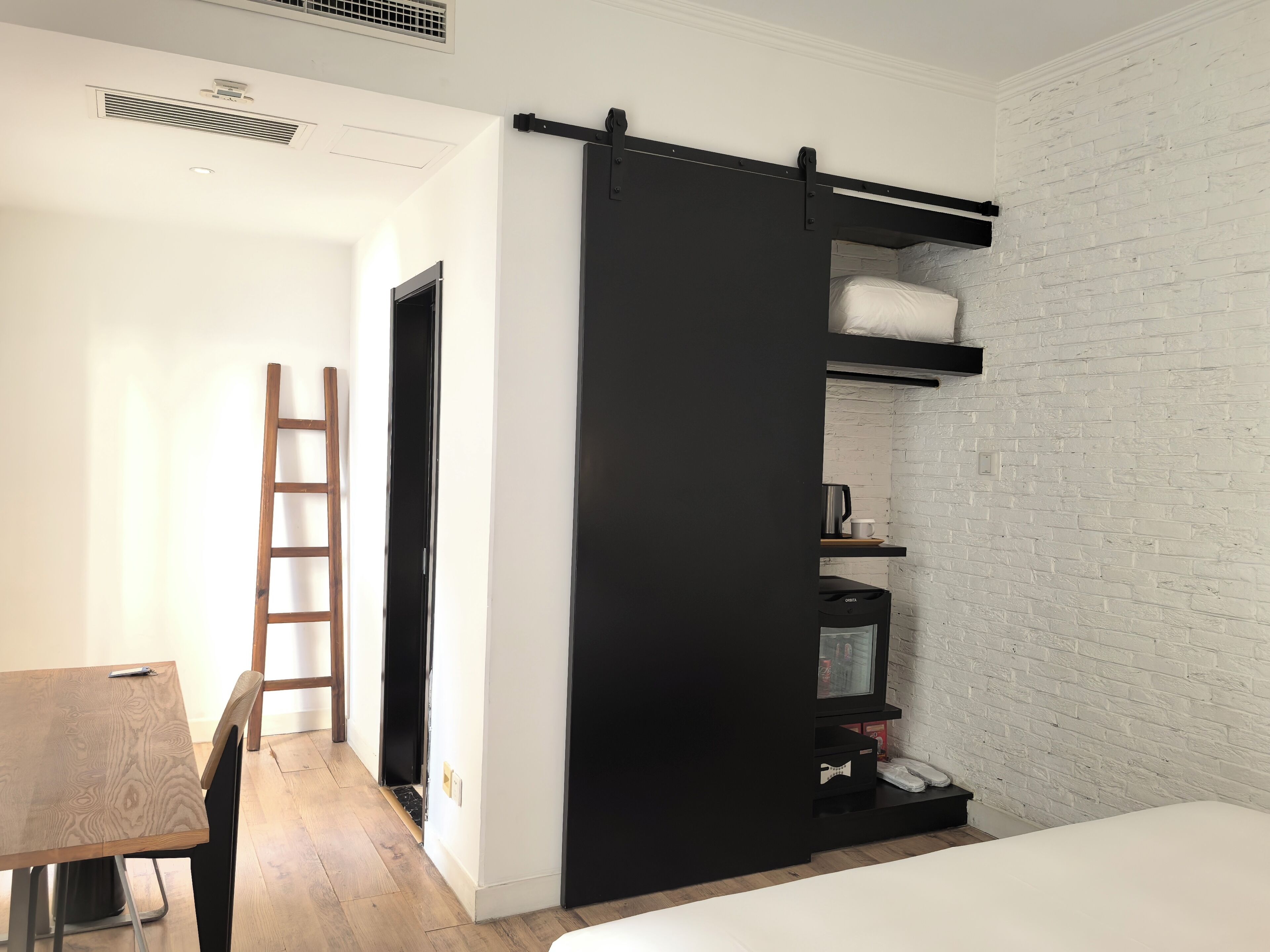 Un dormitorio con cama, armario de puertas correderas, escalera de madera, escritorio y pared de ladrillo blanco.