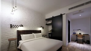 Quarto com cama, escrivaninha e cadeira. Há duas mesas de cabeceira, uma com abajur e outra com mesinha. O quarto tem parede de tijolos brancos e piso de madeira.