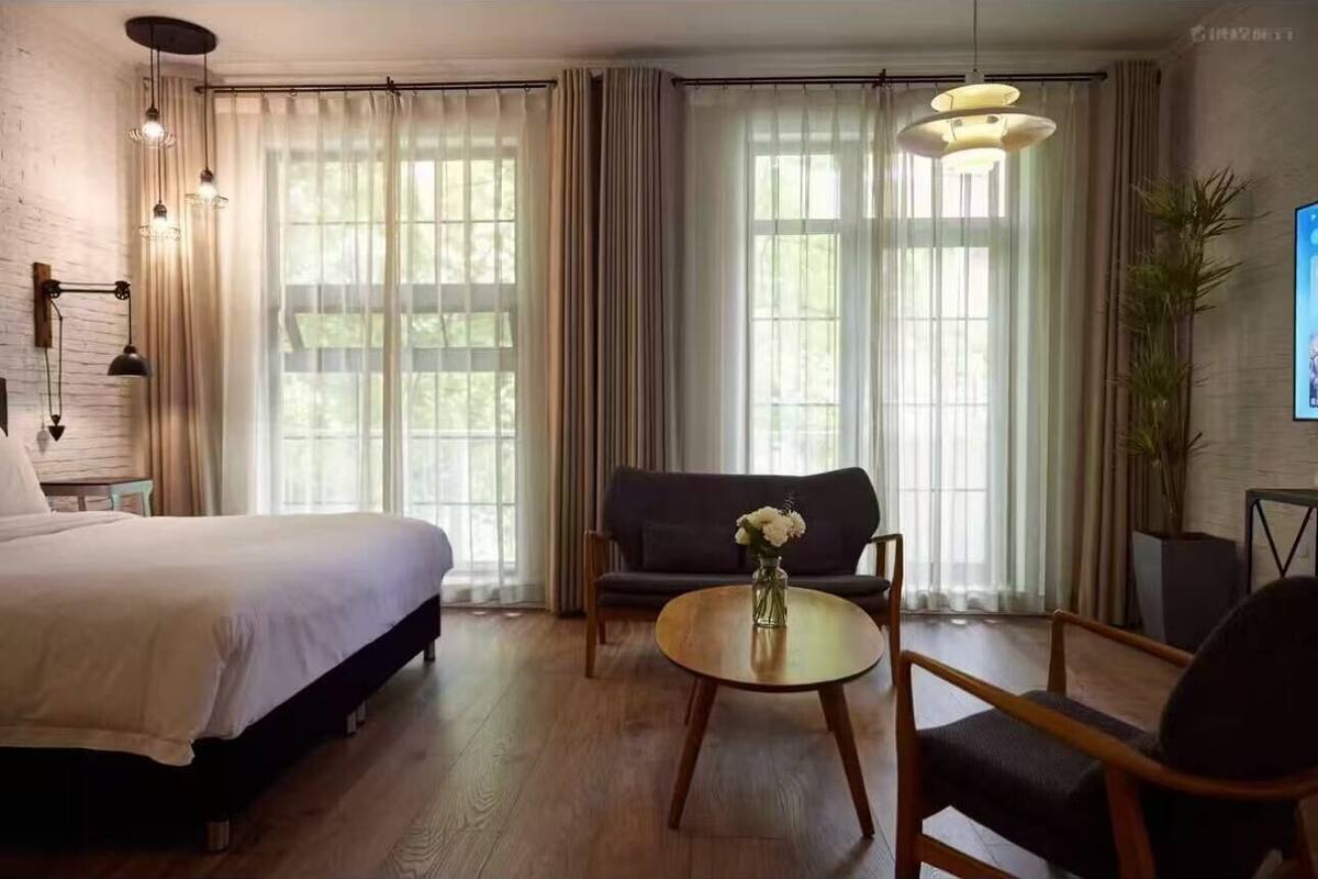 Habitación de hotel con cama, sofá, una mesa redonda con un florero y un ventanal con cortinas.