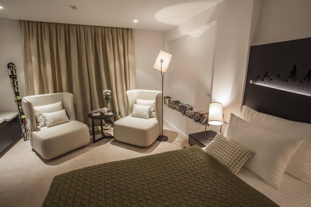 A hotel room with a bed, two armchairs, a small table with a lamp, a bookshelf, and a large window with curtains.