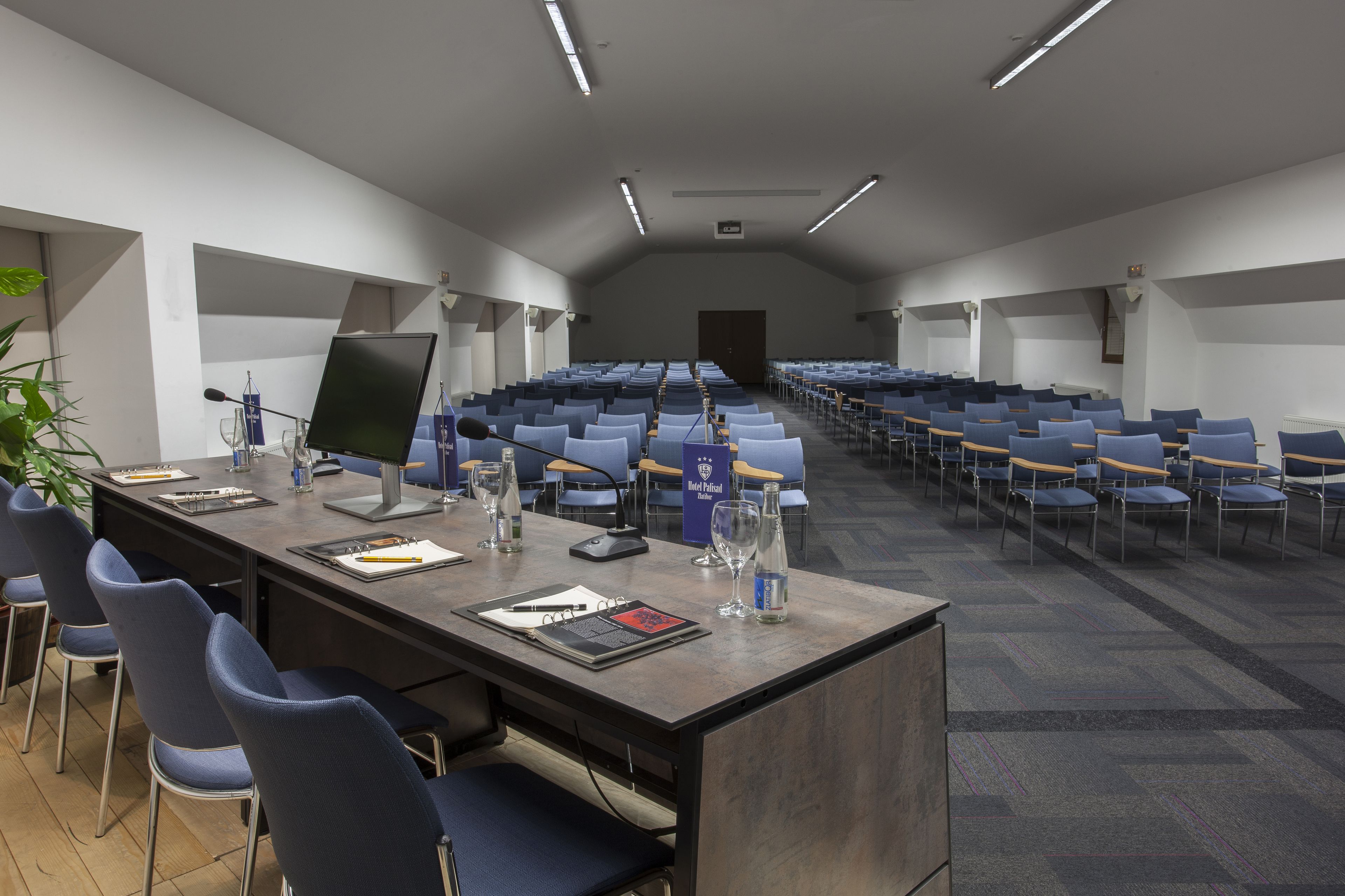 Een congreszaal met rijen blauwe stoelen, een lange tafel met een laptop, glazen en een waterfles.