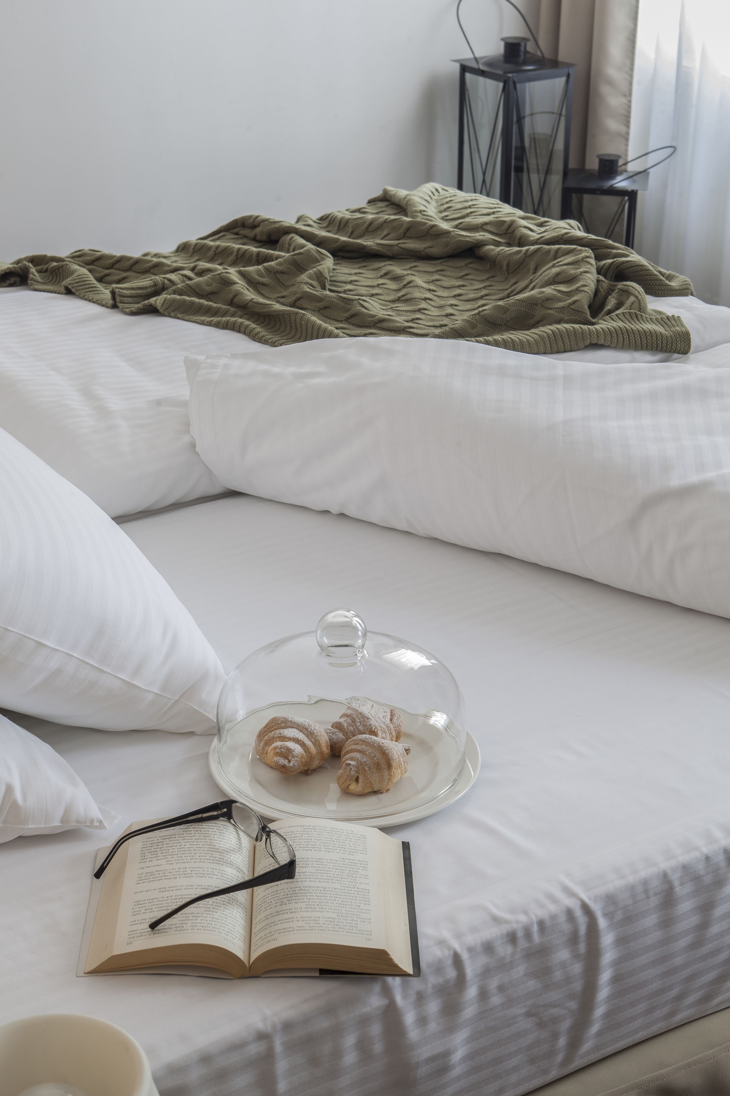 A neatly made bed with white linens, a green blanket, a glass dome with pastries, an open book, and a cup.