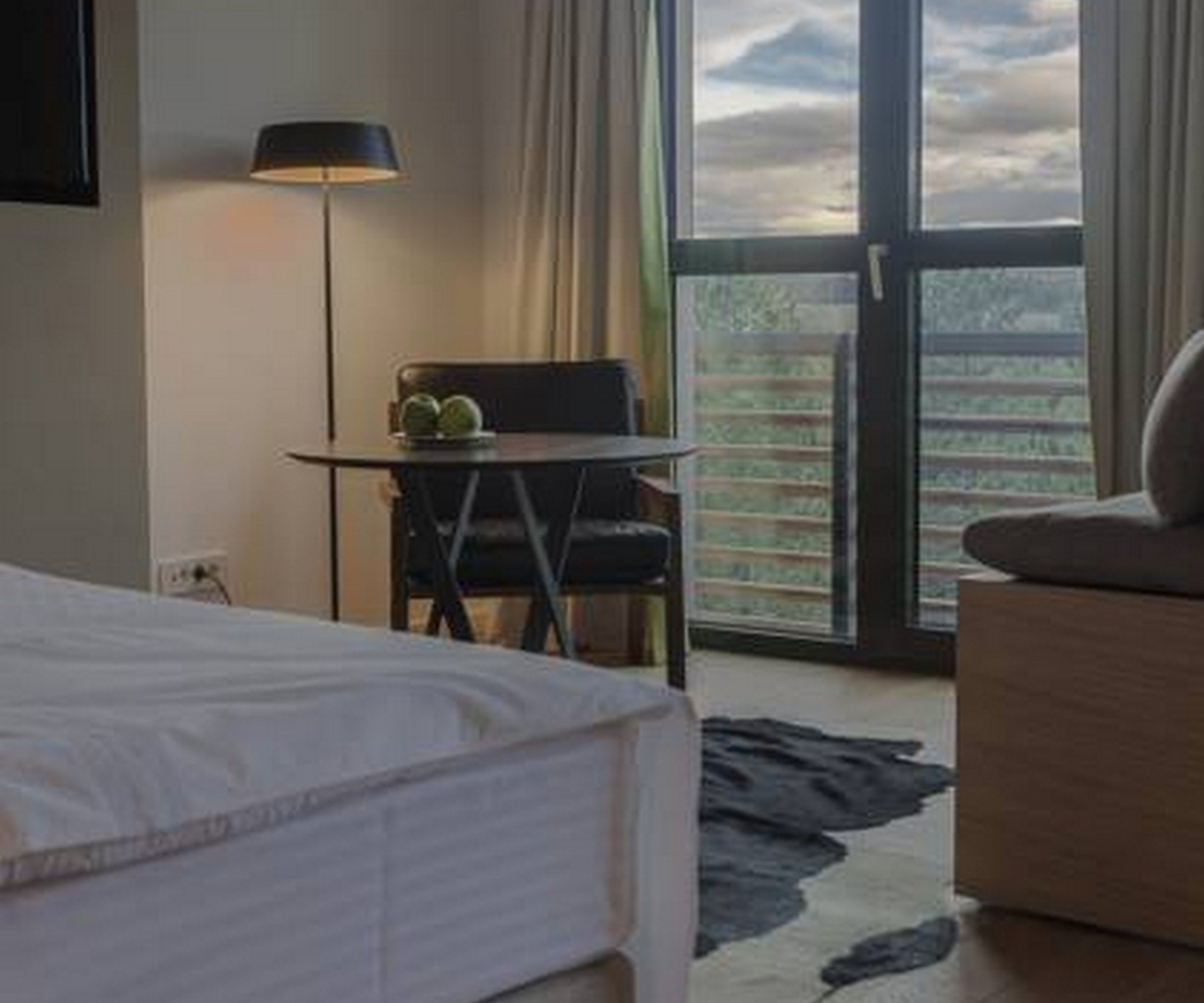 A hotel room with a bed, a desk with a chair, a lamp, and a view of the outdoors through a large window.