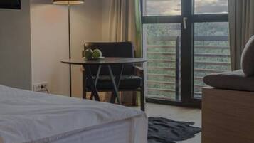 A hotel room with a bed, a desk with a chair, a lamp, and a view of the outdoors through a large window.