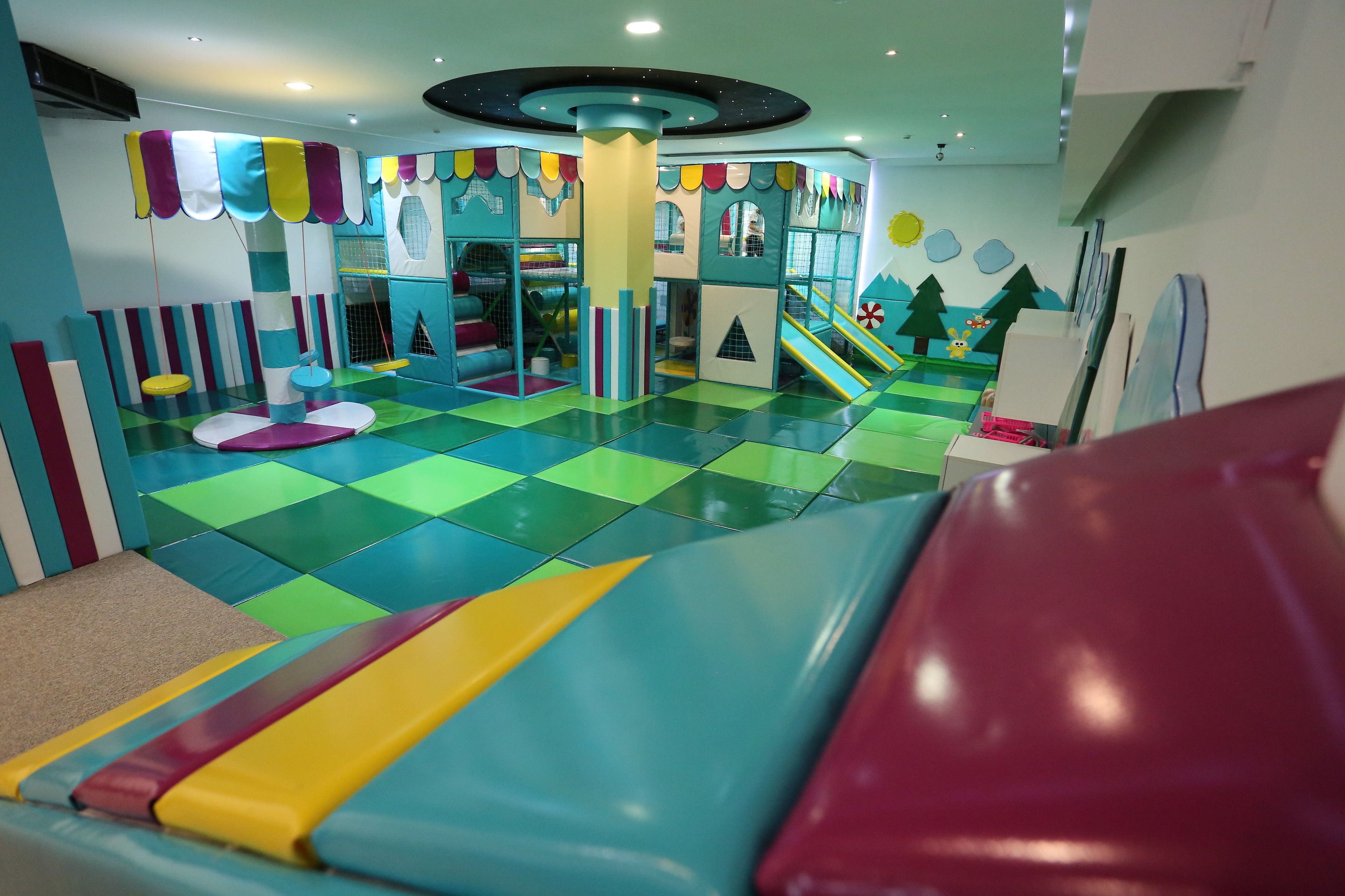 Un espace de jeux pour enfants avec un sol coloré et rembourré, des structures de jeu et un toboggan.