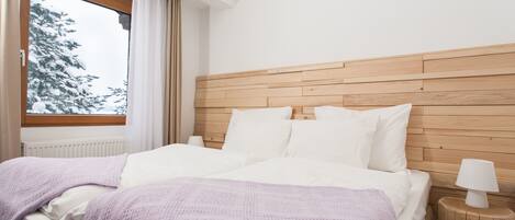 A bedroom with a wooden headboard, white bedding, a window with a view of snow-covered trees, and a bedside lamp on a wooden nightstand.