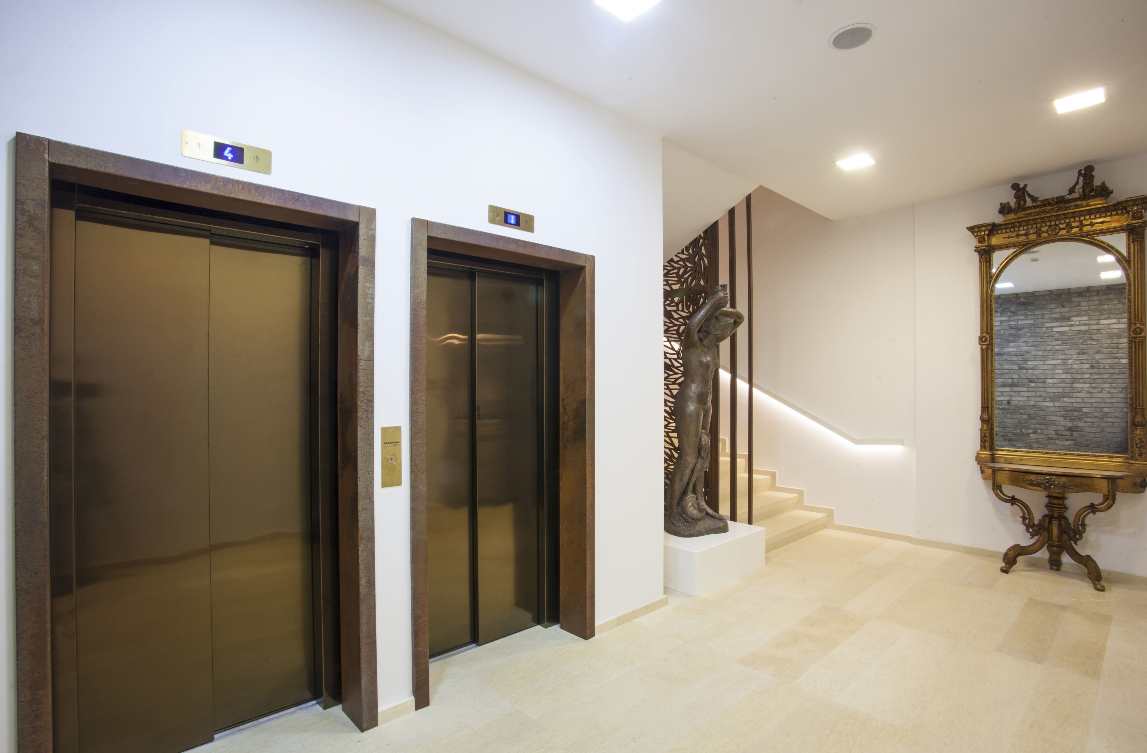 Um elevador moderno com visor digital, escadaria com guarda-corpo metálico decorativo e um espelho grande e ornamentado na parede.