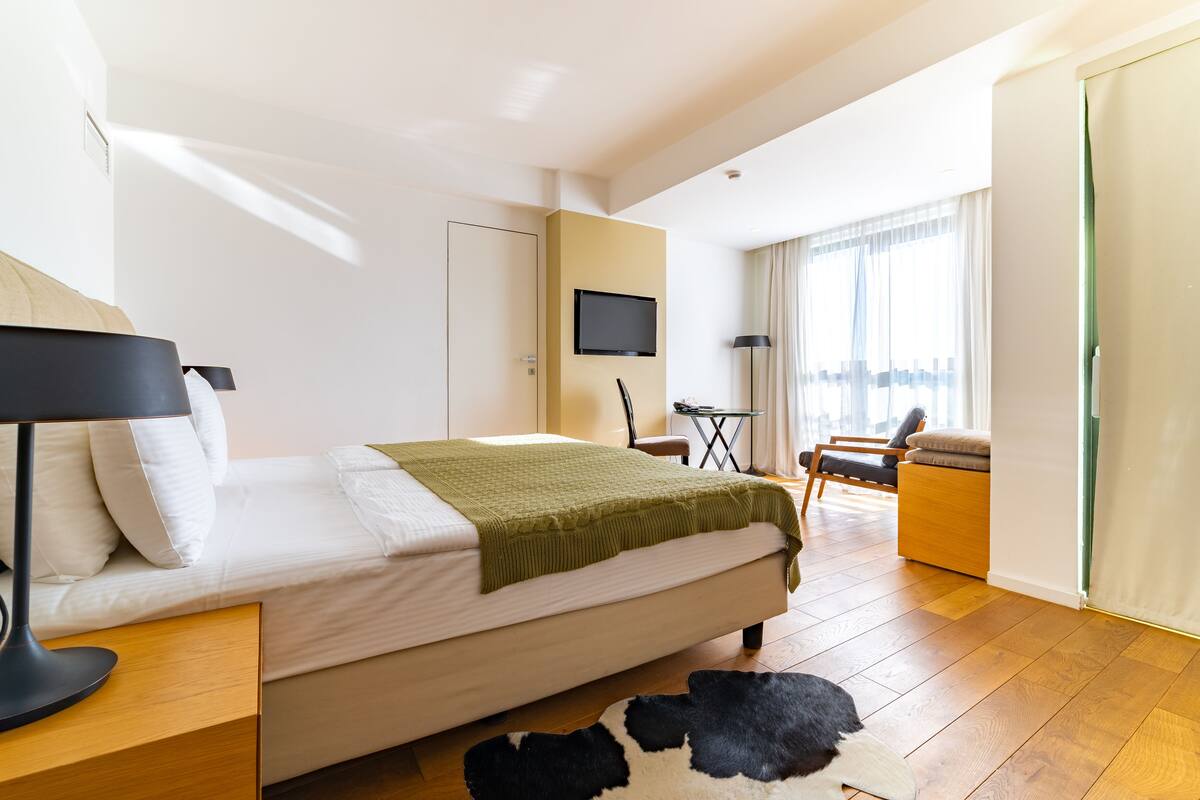 A modern hotel room with a bed, a desk, a TV, and a view of the city.
