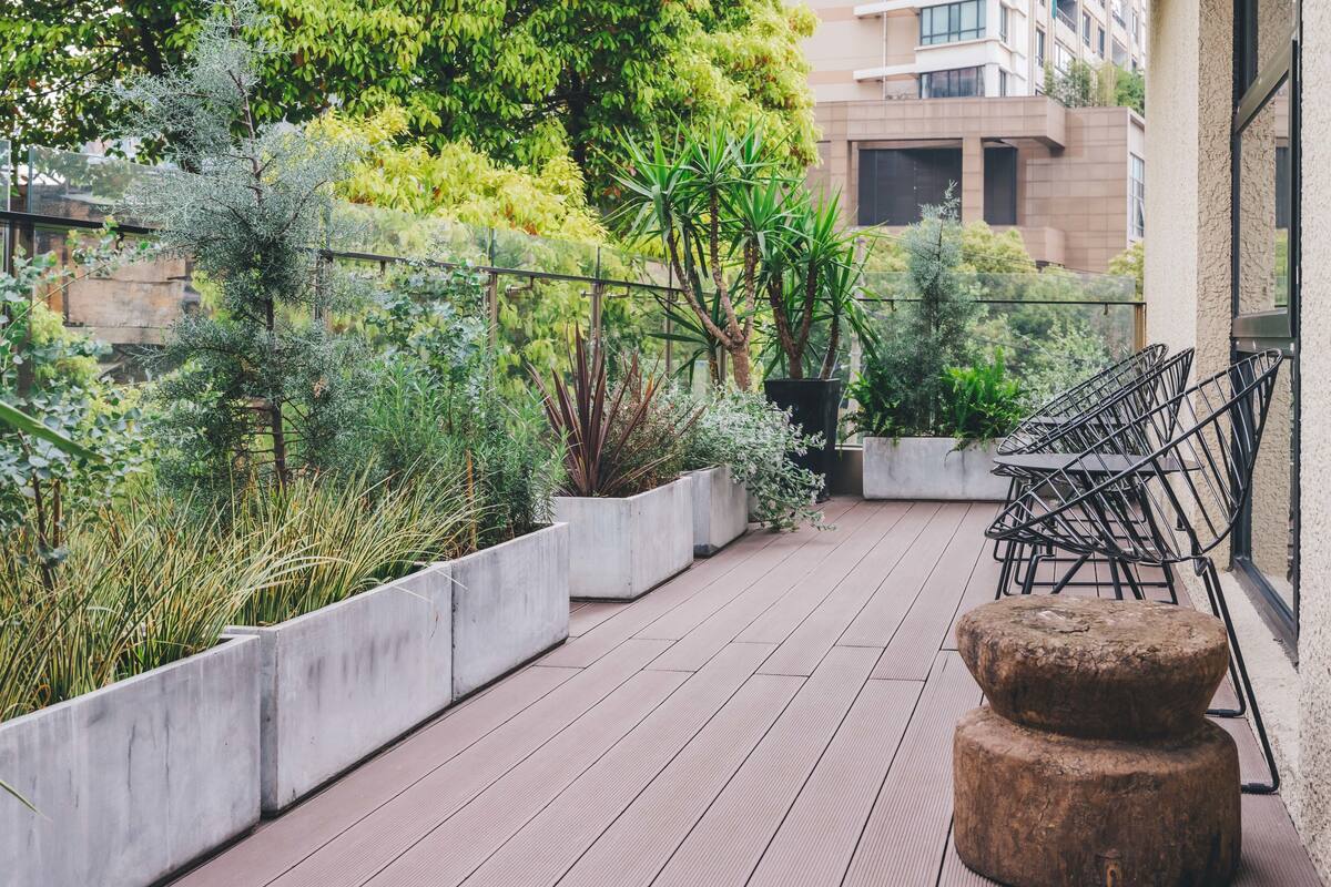 A rooftop garden with concrete planters, a wooden deck, and metal chairs.
