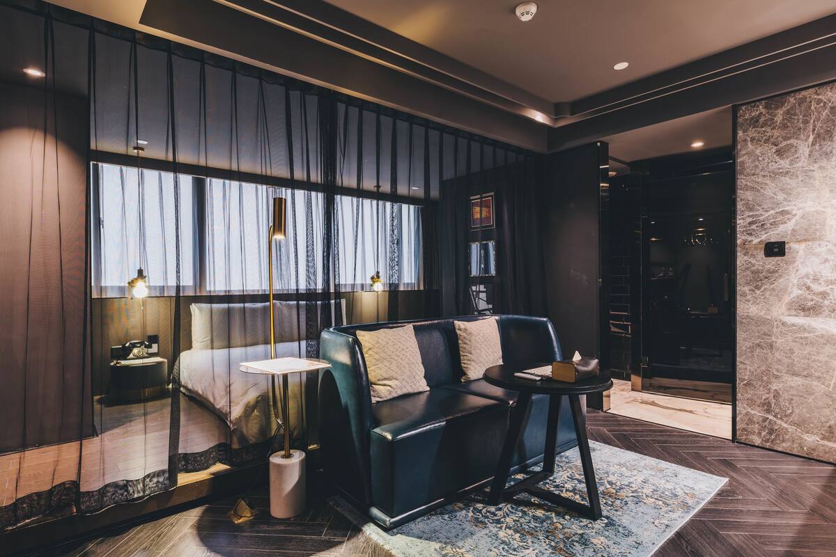 A modern hotel room with a large bed, a sofa, a small table, and a large window with curtains.