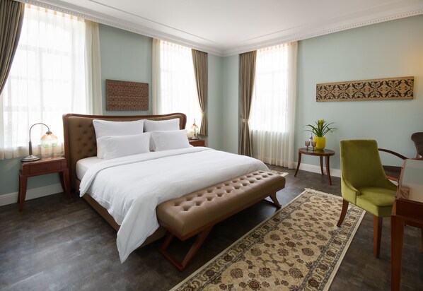 Una habitación de hotel con una cama grande, un banco, un escritorio, una silla, una mesita, una lámpara y una planta en maceta.