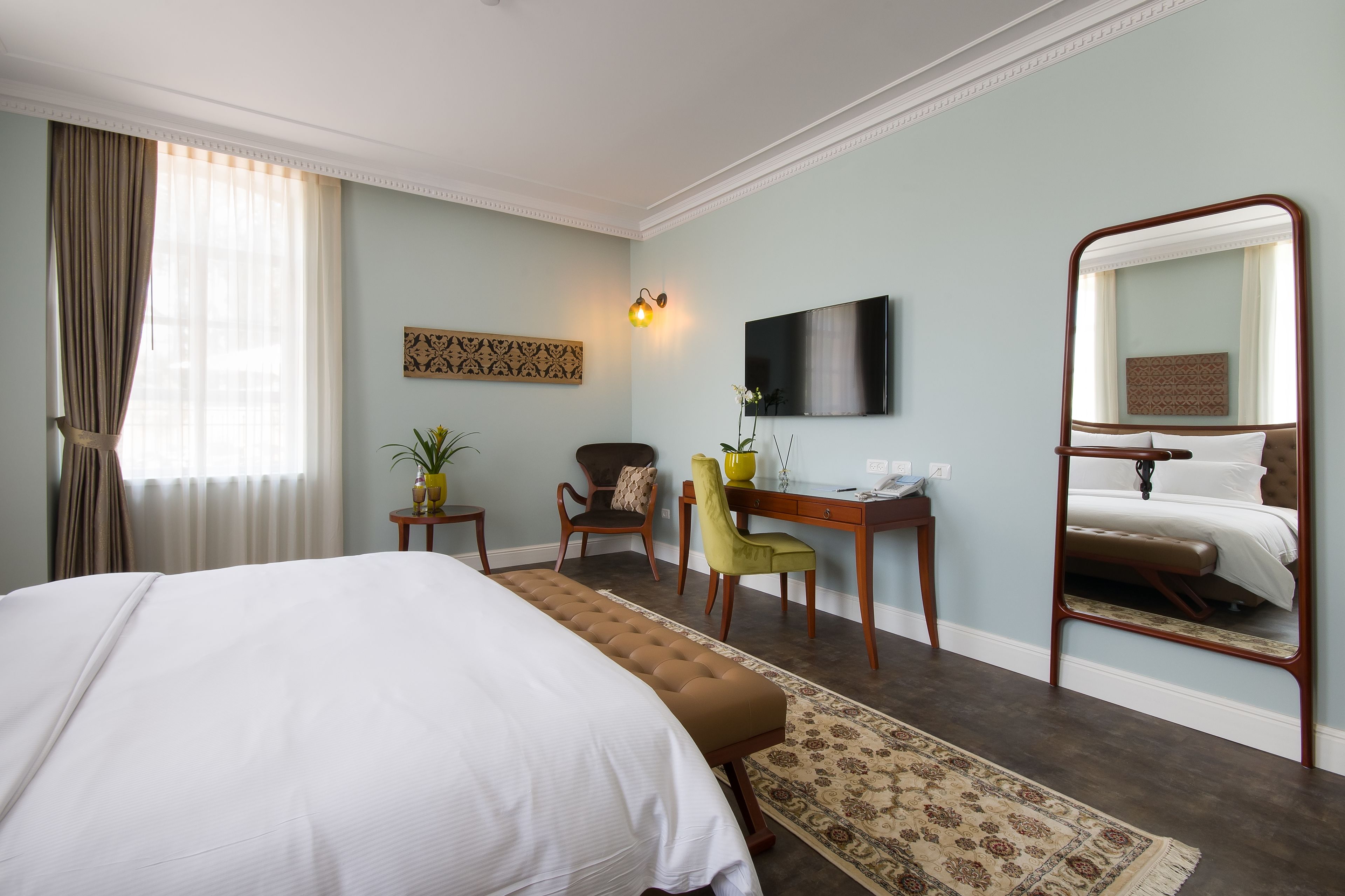 Ein Hotelzimmer mit einem großen Bett, einem Schreibtisch, einem Sessel, einem Spiegel und einem an der Wand befestigten Fernseher.