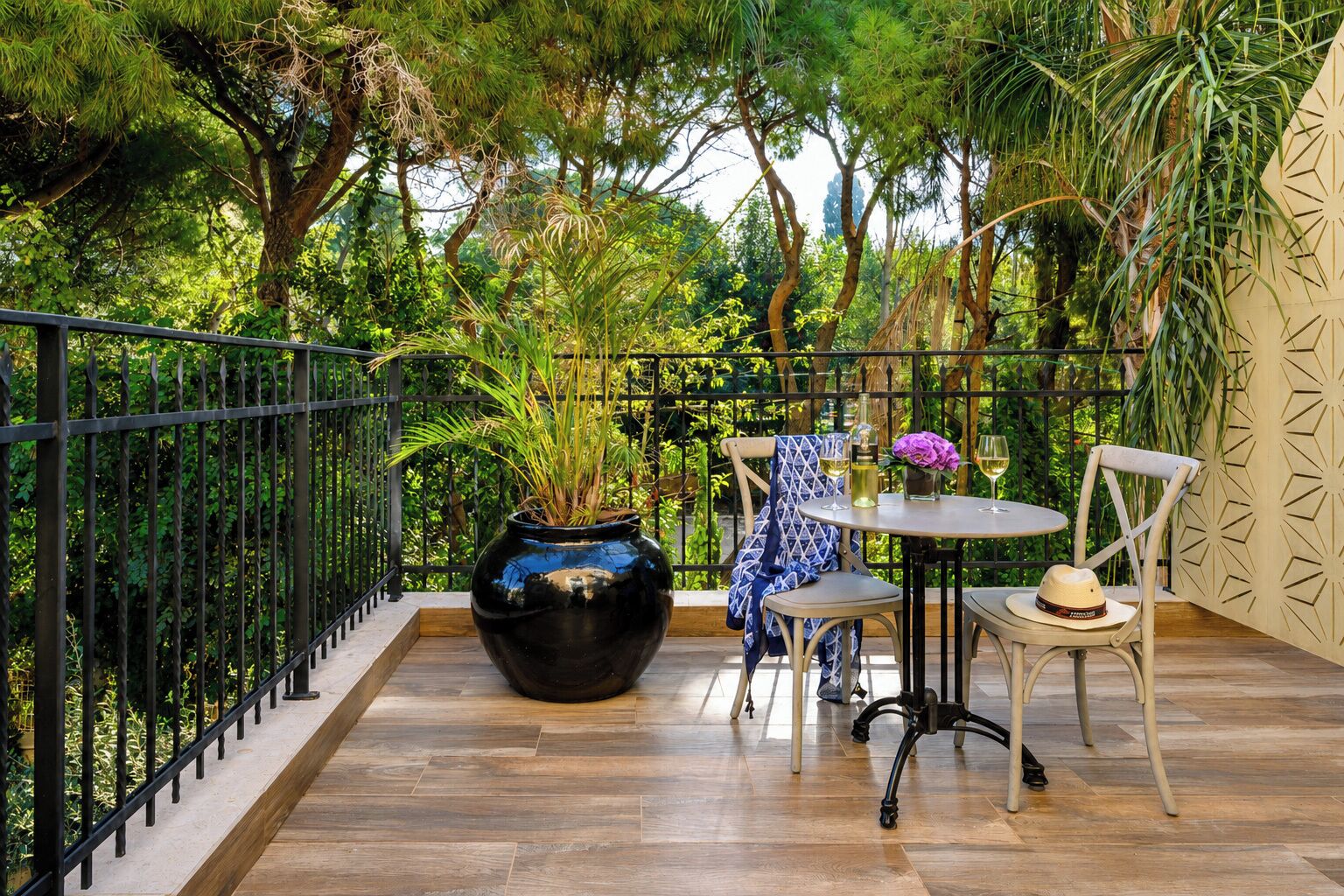 A balcony with a wooden floor, metal railing, a round table with chairs, a vase with flowers, and a potted plant.